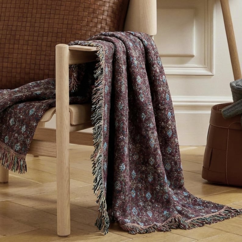 May include: A burgundy throw blanket with a floral pattern and fringed edges is draped over a wooden chair. The blanket features small blue and white floral accents. The chair is made of light-colored wood, and the background includes a leather pillow.