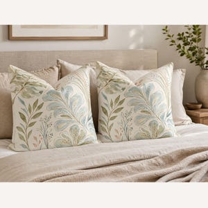 May include: Two square scatter cushions featuring a watercolour-style botanical print on an off-white background. The design displays large, flowing blue and green leaves, delicate stems, and small dotted accents in soft earth tones across the fabric.