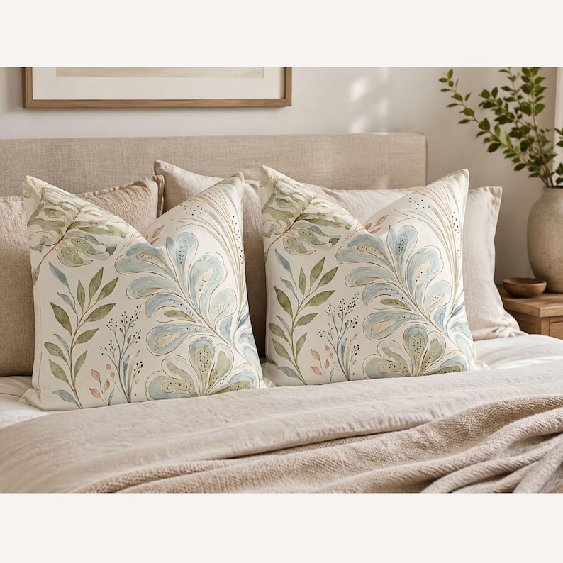 May include: Two square scatter cushions featuring a watercolour-style botanical print on an off-white background. The design displays large, flowing blue and green leaves, delicate stems, and small dotted accents in soft earth tones across the fabric.