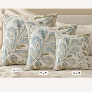 May include: Three square throw pillows featuring a watercolour-style botanical pattern in shades of blue, green, and soft pink on an off-white background. Labels indicate sizes of 28 x 28 cm, 26 x 26 cm, and 24 x 24 cm respectively.
