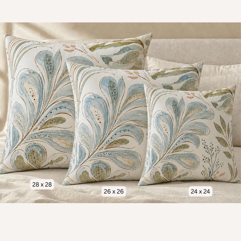 May include: Three square throw pillows featuring a watercolour-style botanical pattern in shades of blue, green, and soft pink on an off-white background. Labels indicate sizes of 28 x 28 cm, 26 x 26 cm, and 24 x 24 cm respectively.