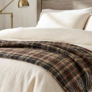 Puede incluir: Una manta tejida con un patr&oacute;n de cuadros en tonos verde oscuro, marr&oacute;n y rojo, colocada sobre una cama con ropa de cama color crema. La manta tiene un acabado texturizado e incluye detalles de flecos en el borde inferior.