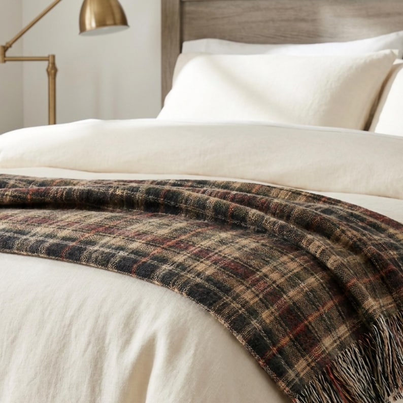 Puede incluir: Una manta tejida con un patr&oacute;n de cuadros en tonos verde oscuro, marr&oacute;n y rojo, colocada sobre una cama con ropa de cama color crema. La manta tiene un acabado texturizado e incluye detalles de flecos en el borde inferior.