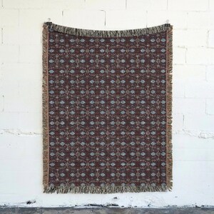 May include: A rectangular throw blanket with a repeating pattern of blue and beige floral designs on a dark brown background. The blanket has a fringed edge and is hanging against a white brick wall.