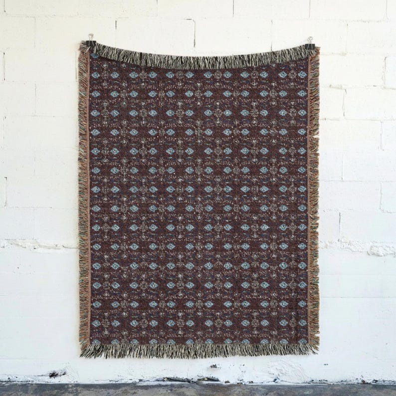 May include: A rectangular throw blanket with a repeating pattern of blue and beige floral designs on a dark brown background. The blanket has a fringed edge and is hanging against a white brick wall.