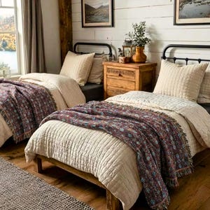 May include: Two twin beds with black metal frames are in a rustic bedroom. Each bed has a beige quilted comforter and pillows, and a patterned throw blanket in shades of burgundy, blue, and brown. A wooden nightstand sits between the beds.