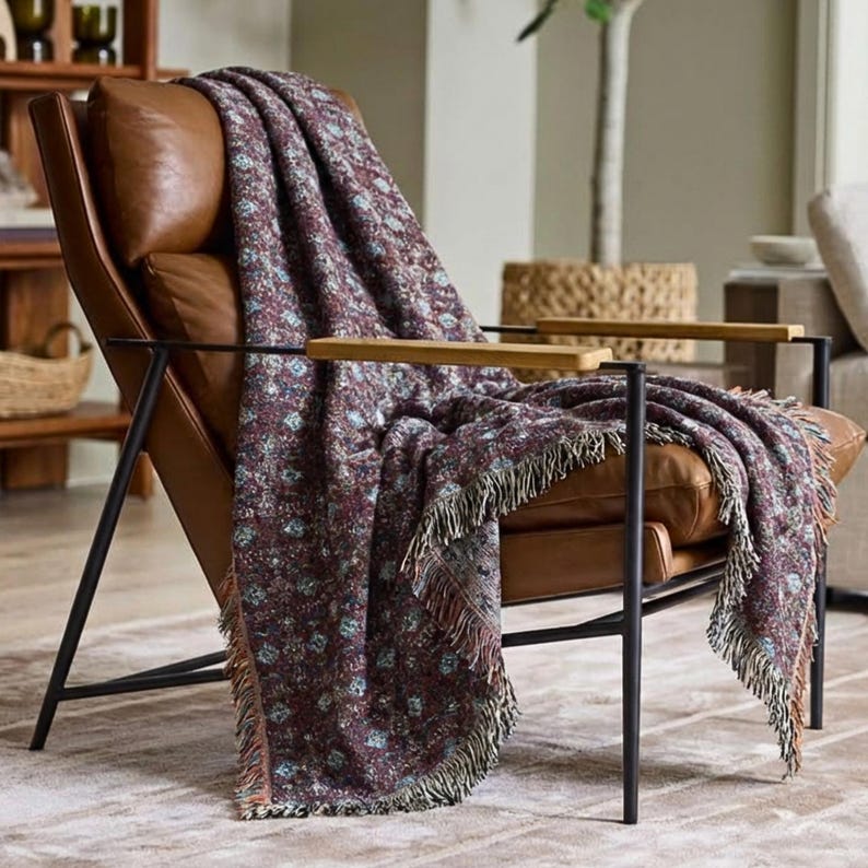 May include: A brown leather armchair with a floral patterned throw blanket. The blanket is primarily burgundy with teal and white accents and fringe trim. The chair has a black metal frame and wooden armrests.