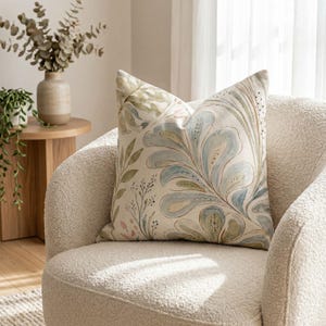 May include: A square throw pillow featuring a watercolour-style botanical print with large, flowing blue and green leaves, delicate stems, and small dotted accents on an off-white background, resting on a textured, cream-coloured armchair.