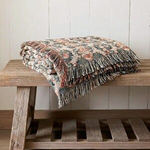 May include: A folded floral patterned throw blanket with fringe. The blanket features a cream background with a repeating pattern of red, green, and brown floral elements. The blanket is resting on a rustic wooden bench.