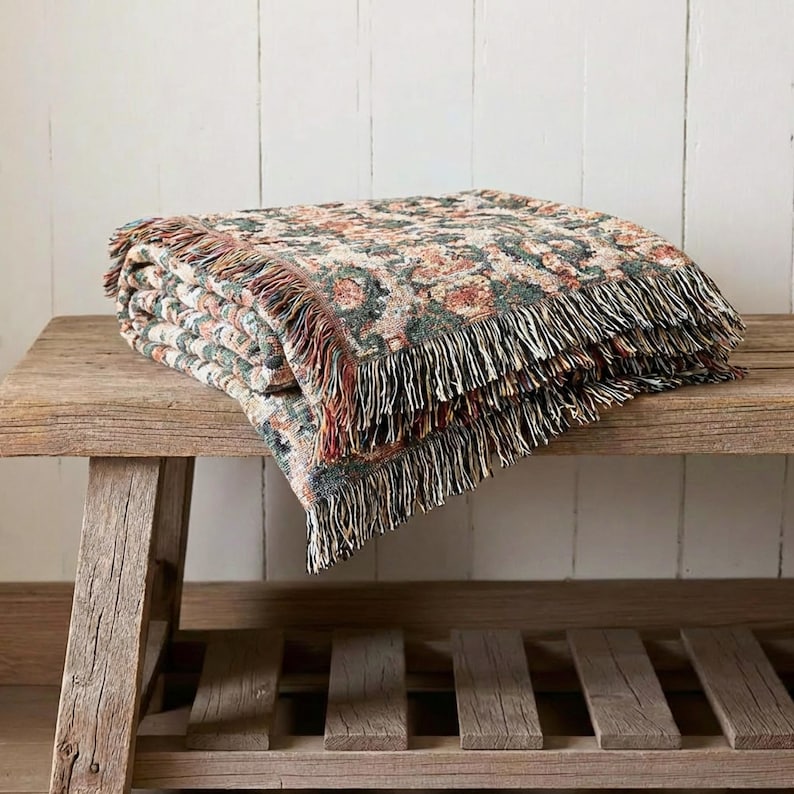 May include: A folded floral patterned throw blanket with fringe. The blanket features a cream background with a repeating pattern of red, green, and brown floral elements. The blanket is resting on a rustic wooden bench.