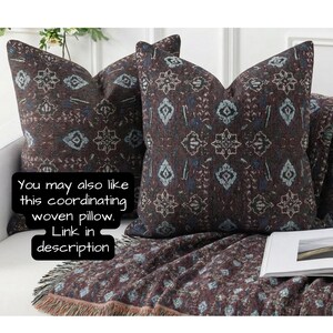 May include: Two decorative throw pillows with a dark brown background and a repeating pattern of white, blue, and teal shapes. A matching throw blanket with fringe is draped over a couch. Text on the image says, "You may also like this coordinating woven pillow. Link in description."