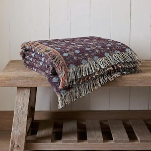 May include: Folded burgundy throw blanket with a repeating floral pattern in blue and cream. The blanket has a fringed edge in shades of cream, red, and brown. The blanket is resting on a rustic wooden bench.