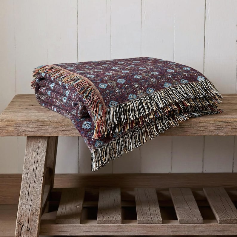May include: Folded burgundy throw blanket with a repeating floral pattern in blue and cream. The blanket has a fringed edge in shades of cream, red, and brown. The blanket is resting on a rustic wooden bench.