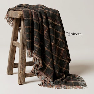 May include: A dark charcoal gray throw blanket featuring a prominent plaid pattern in shades of rust, orange, and cream. The blanket has a fringed edge and is draped over a rustic wooden stool. The text 3sizes is visible on the right side.