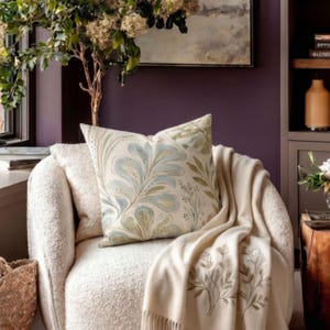 May include: A square throw cushion featuring a cream background with a blue and green botanical leaf pattern, placed on a textured cream armchair draped with a matching cream throw blanket that has embroidered leaf details along the edge.