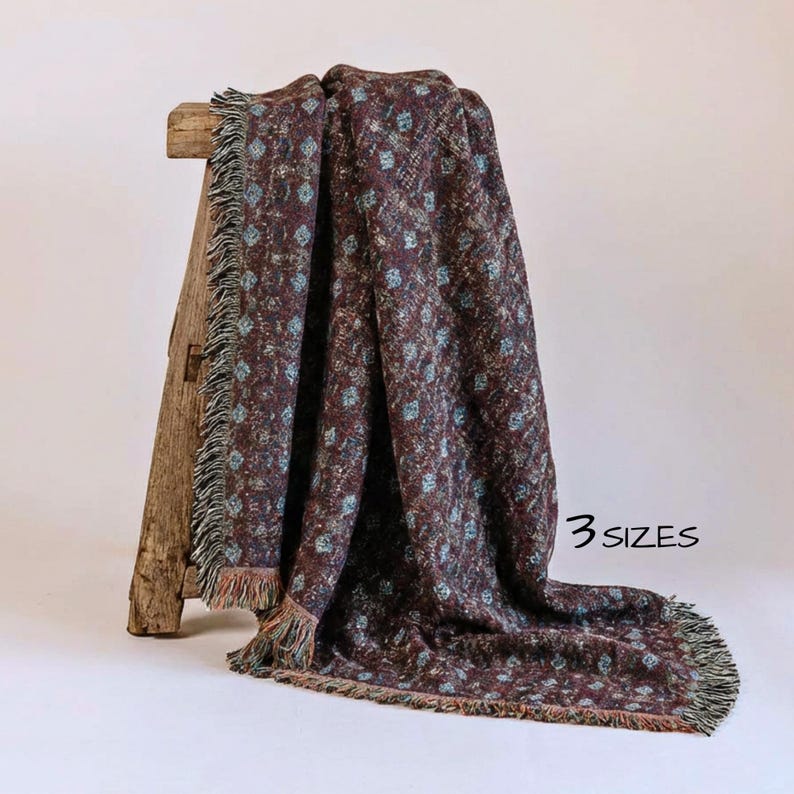 May include: A burgundy throw blanket with a repeating floral pattern in light blue and white. The blanket has a fringed edge and is draped over a wooden stool. The text "3 sizes" is visible.