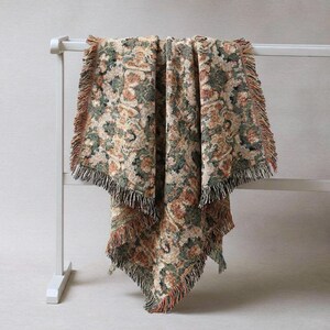 May include: A woven throw blanket with a floral pattern in shades of green, brown, and beige. The blanket has a fringed edge and is draped over a white rack. The overall aesthetic is vintage and cozy, suitable for home decor.