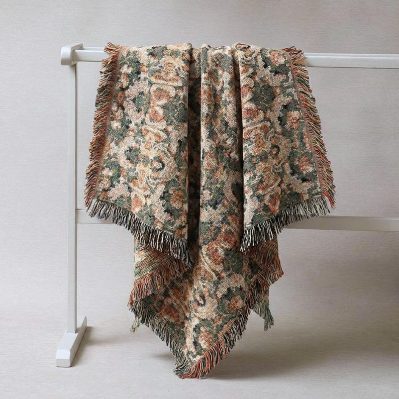 May include: A woven throw blanket with a floral pattern in shades of green, brown, and beige. The blanket has a fringed edge and is draped over a white rack. The overall aesthetic is vintage and cozy, suitable for home decor.