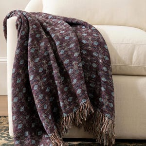 May include: A dark burgundy throw blanket with a repeating pattern of small, light blue shapes. The blanket has a fringed edge and is draped over the arm of a cream-colored armchair. The blanket's texture appears soft and inviting.