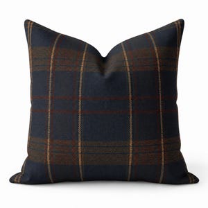 May include: A square throw pillow featuring a dark navy blue base with a woven plaid pattern in shades of rust orange and muted brown. The fabric has a visible textile weave, and the pillow is shaped with a slight indentation at the top center.
