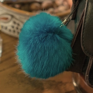 May include: A teal blue faux fur pom pom keychain attached to a black leather bag with a gold buckle.