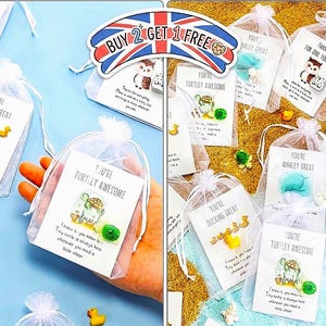 May include: Small gift bags with white organza drawstring closures. Each bag contains a card with a cute animal illustration and a small charm. The cards have phrases like "You're Turtley Awesome" and "You're Whaley Great". A sign reads "Buy 2 Get 1 Free".
