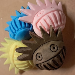 May include: A collection of four sculpted clay figures in pink, blue, yellow, and brown. Each figure features a round shape with spiky hair-like protrusions, two circular eyes, and a wide mouth filled with vertical teeth-like indentations.