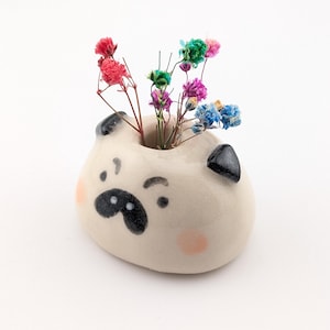 May include: A small, cream-colored ceramic vase shaped like a pug head with black ears, a black nose, and pink cheeks. The vase features a central opening holding a small arrangement of dried flowers in shades of red, blue, green, and purple.