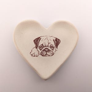 May include: Heart-shaped trinket dish with a brown pug illustration. The dish is off-white and has a slightly textured surface. The pug is depicted in a detailed, sketched style, with its head and front paws visible.