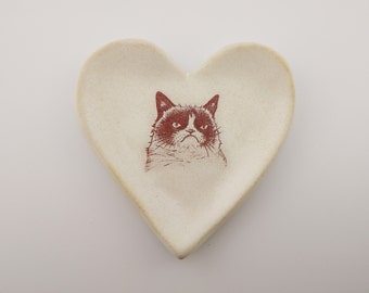 May include: A white ceramic heart-shaped dish with a red and black image of a grumpy cat printed on it.
