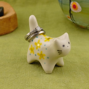 May include: A cream-coloured ceramic cat ring holder featuring a hand-painted pattern of small yellow flowers and light blue dots. The cat has a tall, upright tail designed to hold rings, with two silver-toned rings currently placed on it.