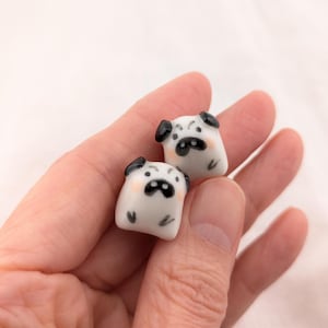 May include: Two small, white ceramic pug figurines with black ears and facial features are held in a hand. The pugs have a cute, cartoonish design, with a black nose and mouth. The background is a soft, off-white color.