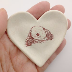 May include: A white ceramic heart-shaped dish with a brown image of a sleeping bunny rabbit.