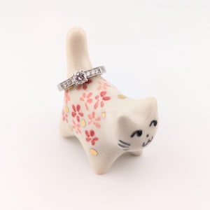 May include: A white ceramic cat ring holder decorated with red floral patterns and gold accents, holding a silver ring with a central pink gemstone and smaller stones. The ring holder is a decorative item.
