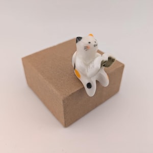 May include: A ceramic cat ornament with white, black, and orange details, sitting on a small brown cardboard box. The cat is in a playful pose. The ornament is a decorative item.