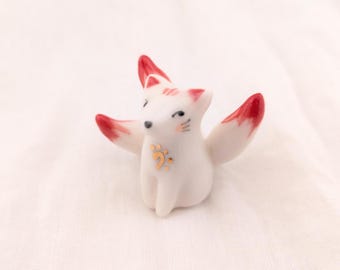 May include: A ceramic fox figurine with white fur and red-tipped tails and ears. The fox has black eyes, pink cheeks, and a gold musical note design on its chest. The figurine is set against a white background.
