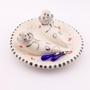 May include: A white ceramic ring dish featuring two sculpted seal figures with black painted details. The dish is decorated with small red heart motifs and a black dashed rim. Two silver-toned rings are placed on the seal heads, and a pair of blue drop earrings rests on the base.