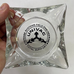 May include: A clear glass ashtray with a star-shaped design. The ashtray has a black and white logo that reads "Defense Systems Field Engineering UNIVAC Total Support".
