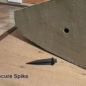 Peut inclure: Un pic de sécurité en plastique noir est posé sur une surface en bois à côté d'un grand objet rectangulaire brun clair. Le texte "Secure Spike" est visible en bas de l'image. L'objet a un trou.