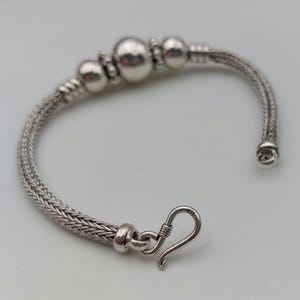 Può includere: Bracciale in argento con un design a catena intrecciata e una chiusura a gancio. Il bracciale presenta tre grandi perle rotonde e perle più piccole. Il gioiello è esposto su una superficie bianca.