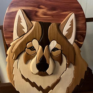 May include: A wooden wall hanging featuring a detailed relief carving of a wolf's face. The artwork uses various shades of natural wood, including light and dark browns, and cream, with a circular background.