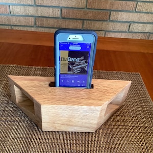 May include: A wooden phone stand with a triangular design, holding a smartphone displaying a "Big Band Radio" interface. The stand is made of light-colored wood and sits on a woven mat.