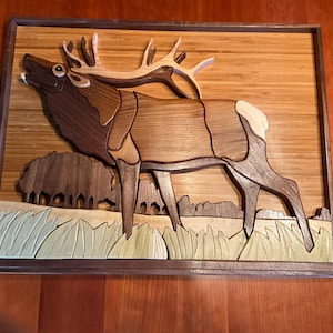 May include: Wooden elk wall art. The artwork features a detailed elk with large antlers, crafted from various shades of wood. The elk is set against a backdrop of trees and grass, all within a framed border. The piece showcases a natural, rustic aesthetic.