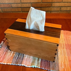 May include: A rectangular wooden tissue box cover featuring a light-colored wood body with a dark wood center panel and visible finger joint construction at the corners. A single white paper tissue extends from the rectangular opening on top.