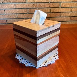 May include: A wooden tissue box with a multi-toned, striped design. The box is square and has a slot on top for tissue dispensing. A white tissue is partially pulled out. The box sits on a white, crocheted doily.