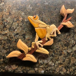 May include: A wooden frog sculpture perched on a branch with leaves. The frog is light yellow and brown, with orange eyes. The branch and leaves are a darker brown and tan. The sculpture is likely handmade.