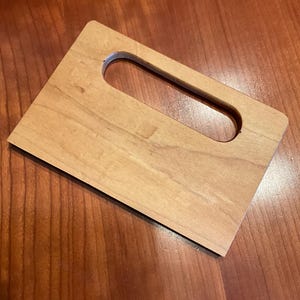 May include: A light-colored wooden cutting board with a rectangular shape and a handle cutout. The board has a smooth surface and is set against a wooden background.