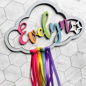 May include: A gray wooden cloud with the name "Evelyn" spelled out in rainbow colors. A white star is on the right side of the cloud. Rainbow colored ribbons hang from the bottom of the cloud.