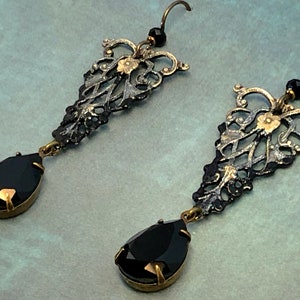 May include: A pair of black and gold dangle earrings with teardrop-shaped black gemstones. The earrings feature intricate filigree designs.