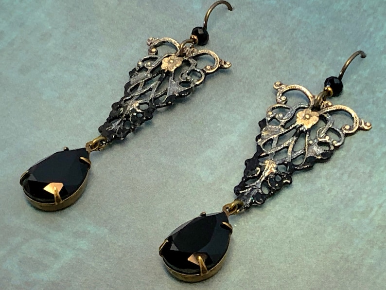 May include: A pair of black and gold dangle earrings with teardrop-shaped black gemstones. The earrings feature intricate filigree designs.
